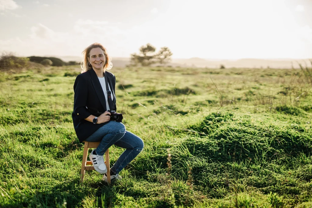 A smiling female photographer sitting on a wooden stool in a sunlit green field, holding a camera. Relaxed and natural outdoor portrait.