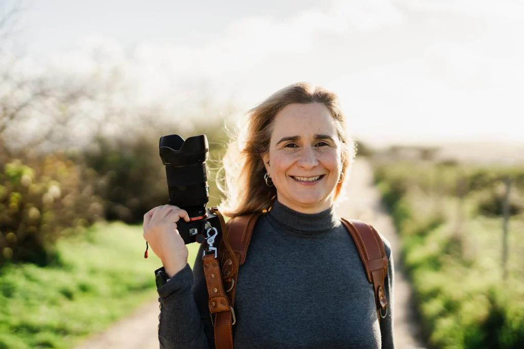 A smiling professional photographer holding a camera over her shoulder while standing on a sunlit path. Dressed in a stylish turtleneck and leather harness, she exudes warmth and confidence, embodying her passion for capturing beautiful moments in Portugal.