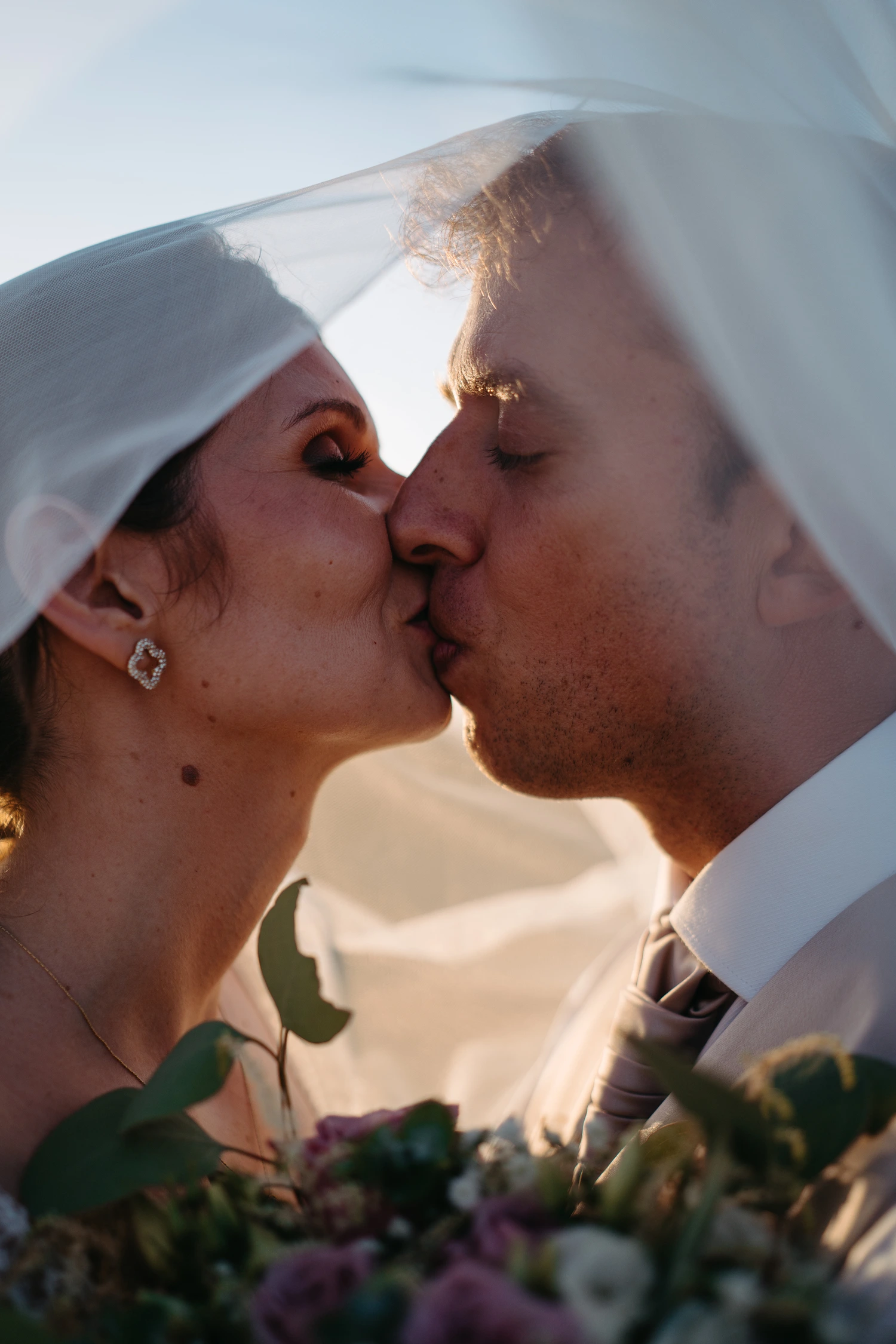 A newlywed couple shares an intimate kiss beneath the bride’s veil during their Lisbon wedding, bathed in warm golden sunlight, capturing a timeless and romantic moment.