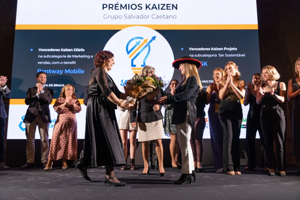 A celebratory awards ceremony with women on stage exchanging flowers and applause under bright event lighting in Lisbon