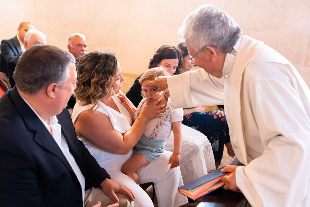 A priest gently blesses a little girl during her baptism as her mother holds her lovingly in a church setting.
