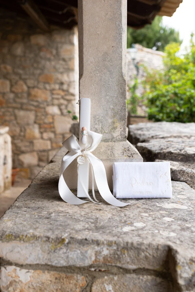A white baptism candle with an elegant ribbon and an embroidered handkerchief placed on a rustic stone ledge.