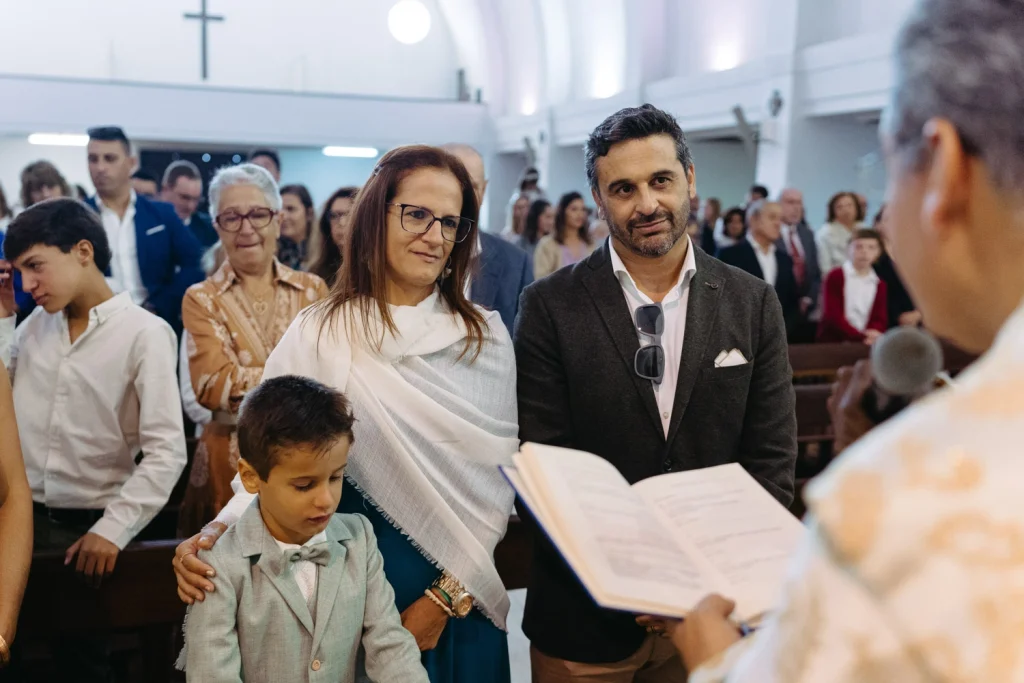 Parents standing with their son during a baptism ceremony in a beautifully lit Portuguese church.