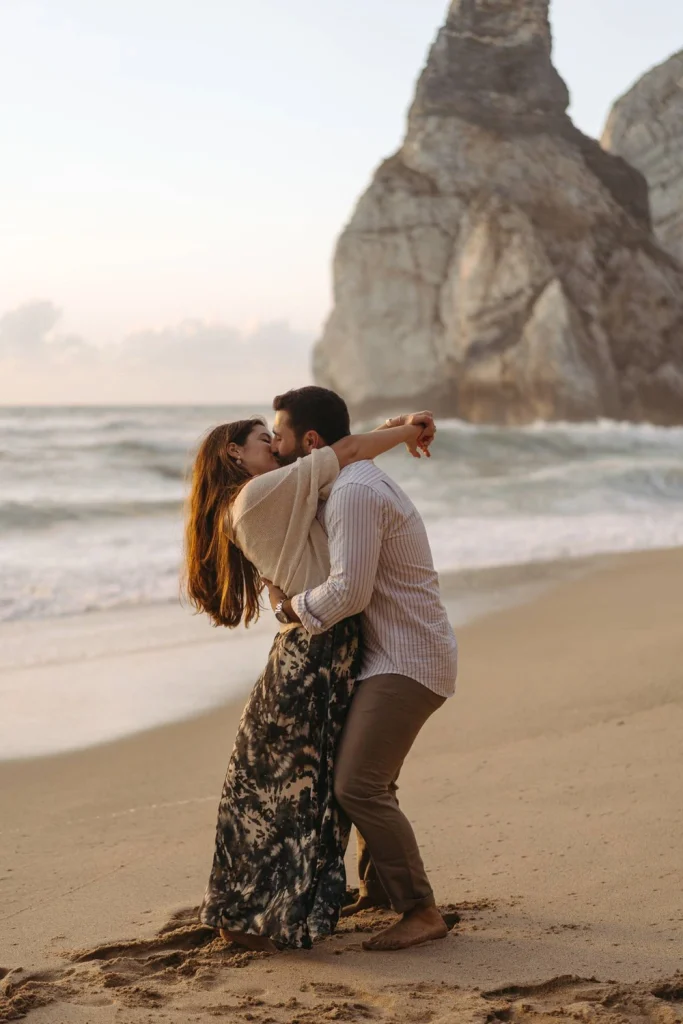 Playful couple enjoying a beach photoshoot in Portugal, surrounded by golden cliffs and foamy waves.
