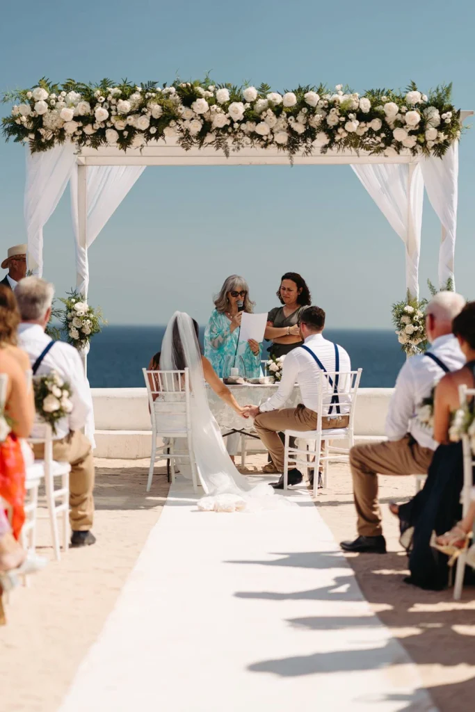 couple hold hands under a floral arch as they exchange vows by the ocean