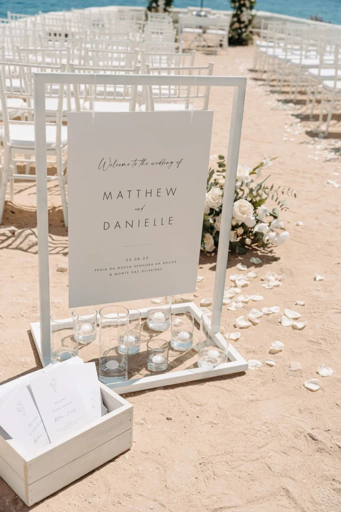 Elegant wedding welcome sign with white flowers and candles on a sandy beach ceremony setup in Algarve