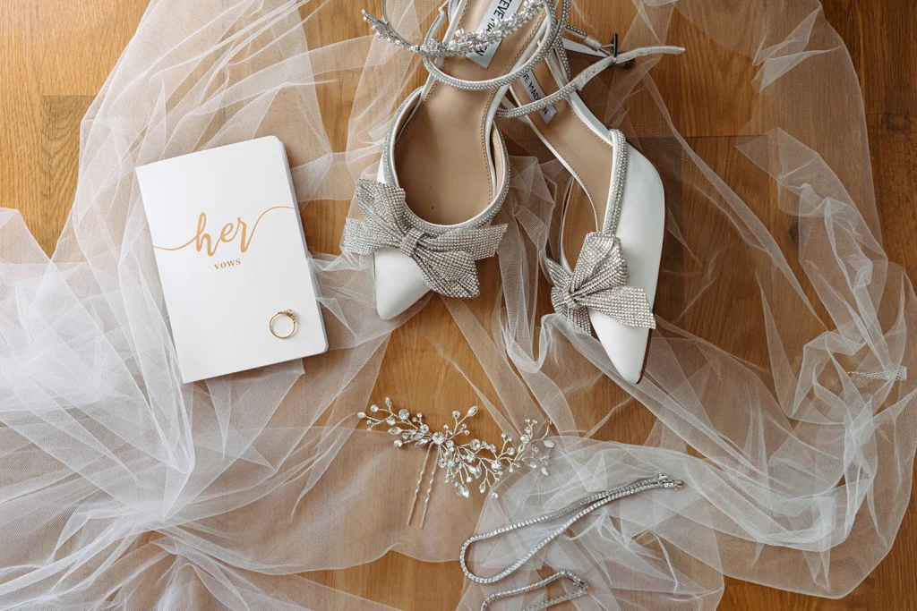 Elegant bridal accessories, including sparkling heels, a vow book, and delicate jewelry, laid out on soft tulle fabric.