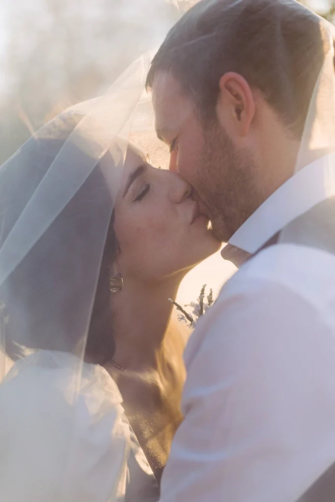 Bride and groom share a romantic kiss under the veil, bathed in soft golden light.
