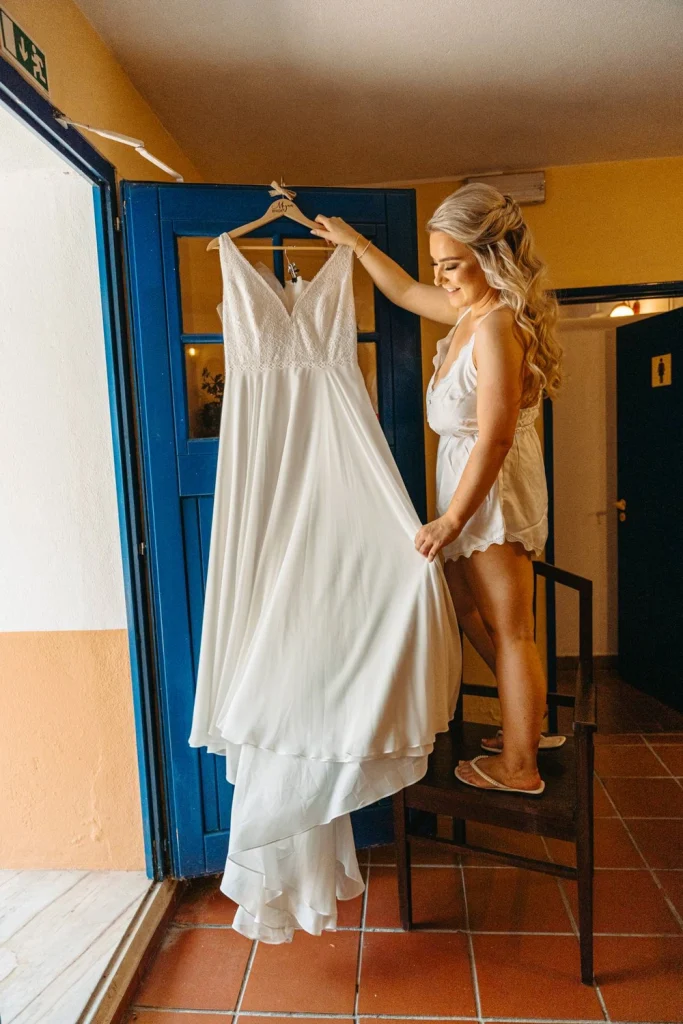 The bride admires her elegant white wedding dress, holding it up against a rustic blue door in Portugal.