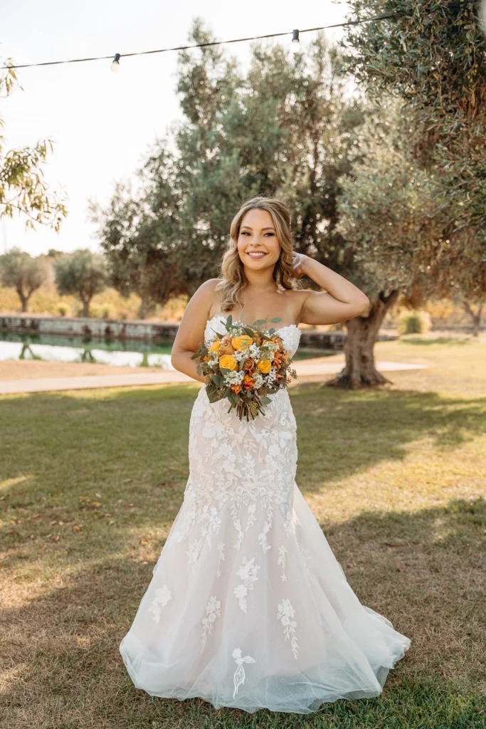 A glowing bride poses outdoors, holding a stunning bouquet of orange and white flowers, with golden sunlight filtering through the trees.