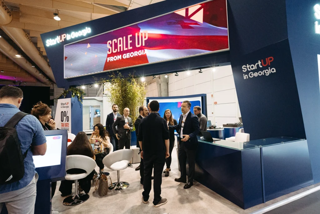 A professional networking event at a modern exhibition booth, where attendees engage in conversations in websummit