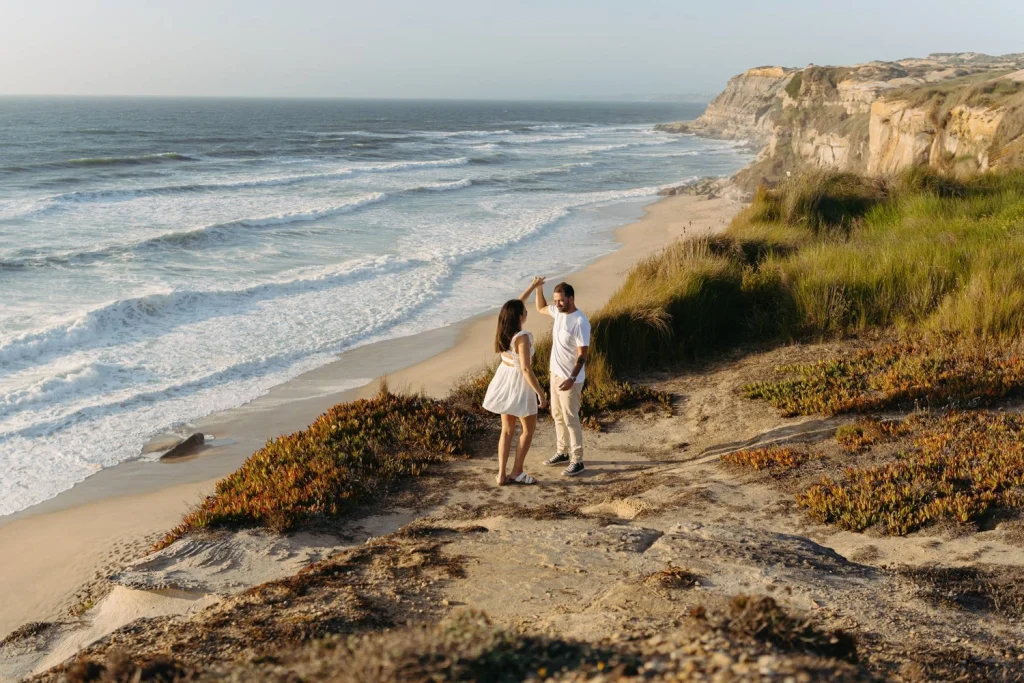 A couple dances on a scenic cliffside overlooking the ocean, with waves crashing below