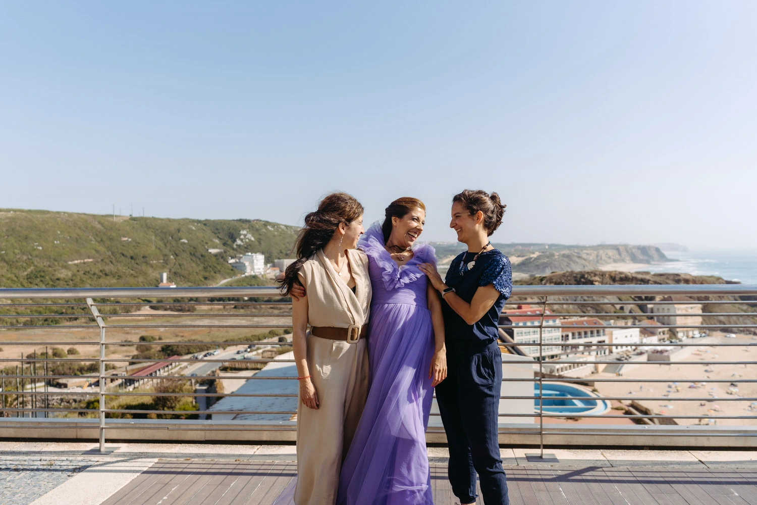 Woman in a flowing lavender gown shares a joyful moment with friends on a scenic terrace overlooking the beach during a birthday celebration in Portugal.