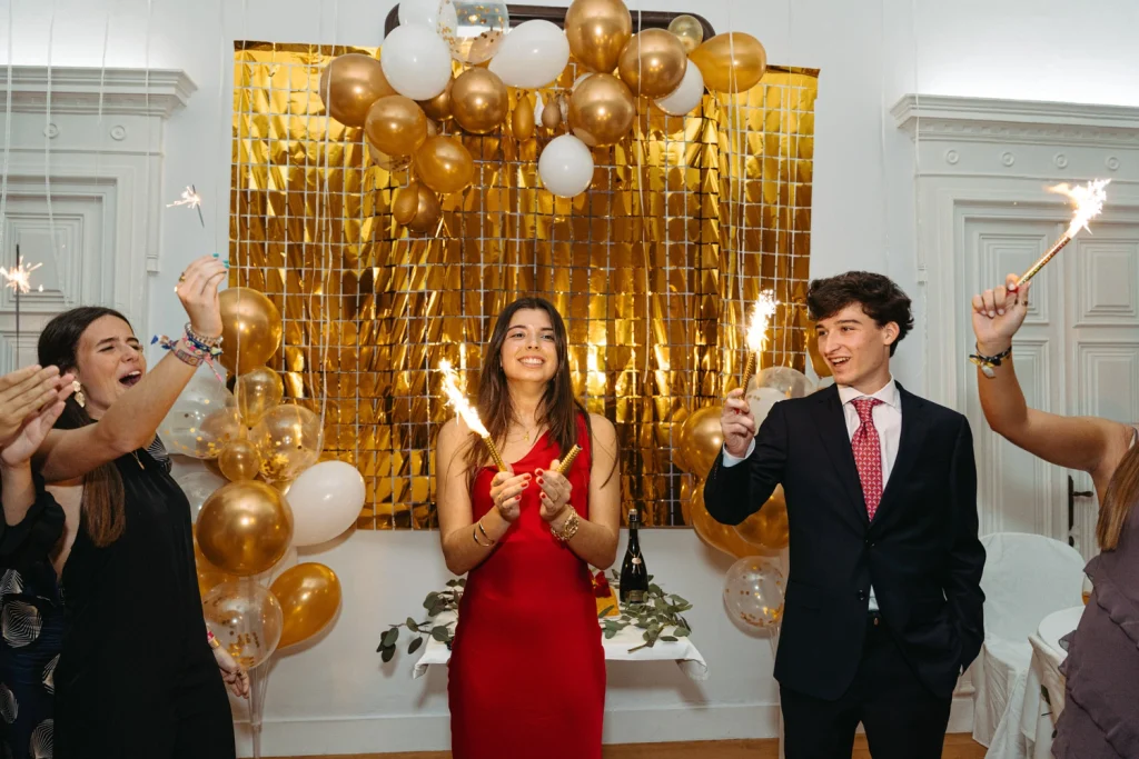 A young woman in a red dress celebrates her birthday with sparklers, surrounded by friends and golden balloons in lisbon