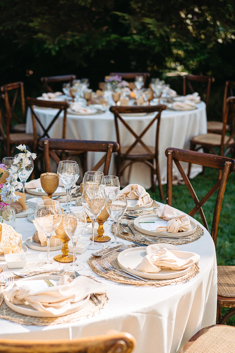 A beautifully set wedding table with gold and white details, fine glassware, and rustic chairs in an outdoor venue.