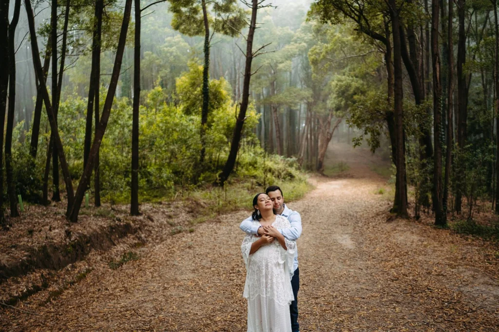 An engaged couple embraces and smiles on a scenic forest road, enjoying a romantic moment.