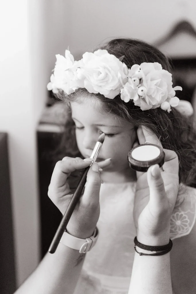 A young flower girl wearing a white floral crown as she gets her makeup done for a baptism.