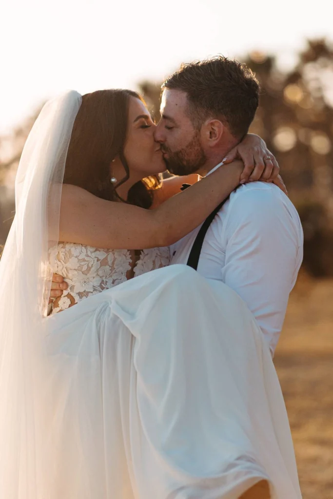 The groom lifts his bride in a joyful embrace as they share a kiss at sunset.