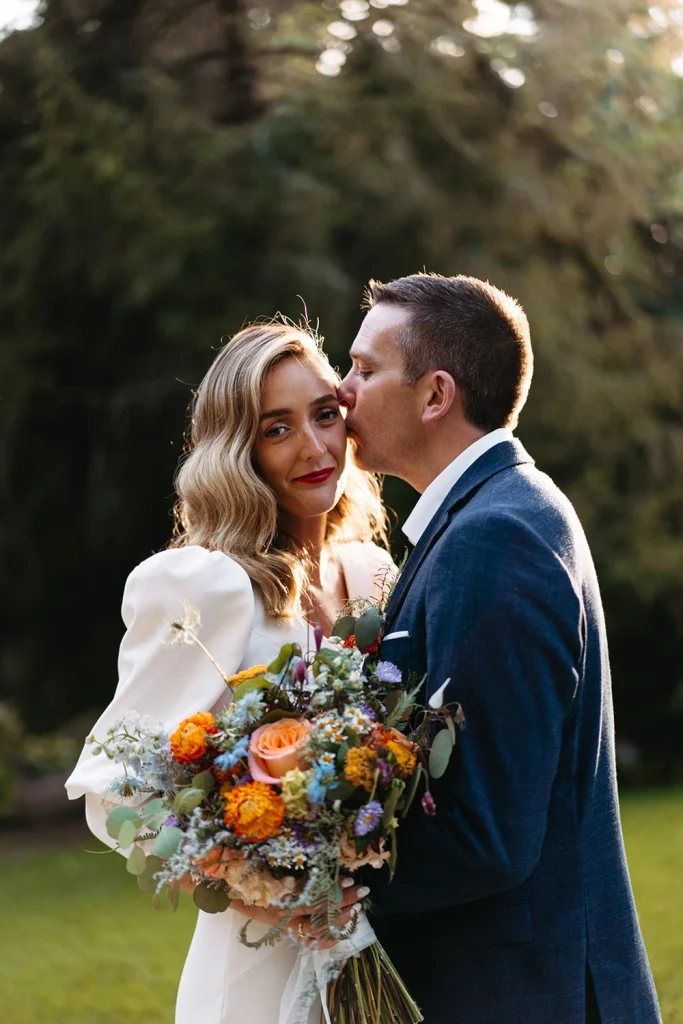 The groom kisses his bride on the forehead in golden evening light, holding her floral bouquet.