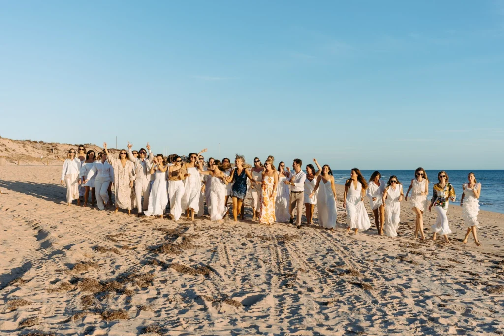 A large group of friends dressed in white, walking together on the beach at sunset, celebrating in style in the beach in algarve