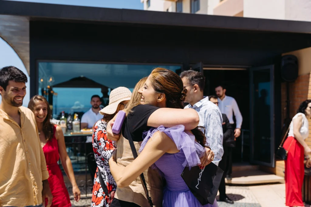 wo women embrace with warm smiles during a daytime event, surrounded by friends in a lively outdoor setting in ericeira