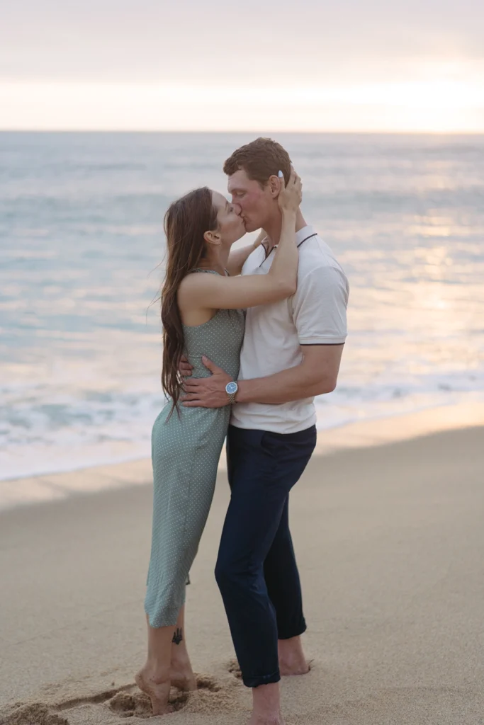 A couple sharing a romantic kiss by the ocean as the sun sets in the background.