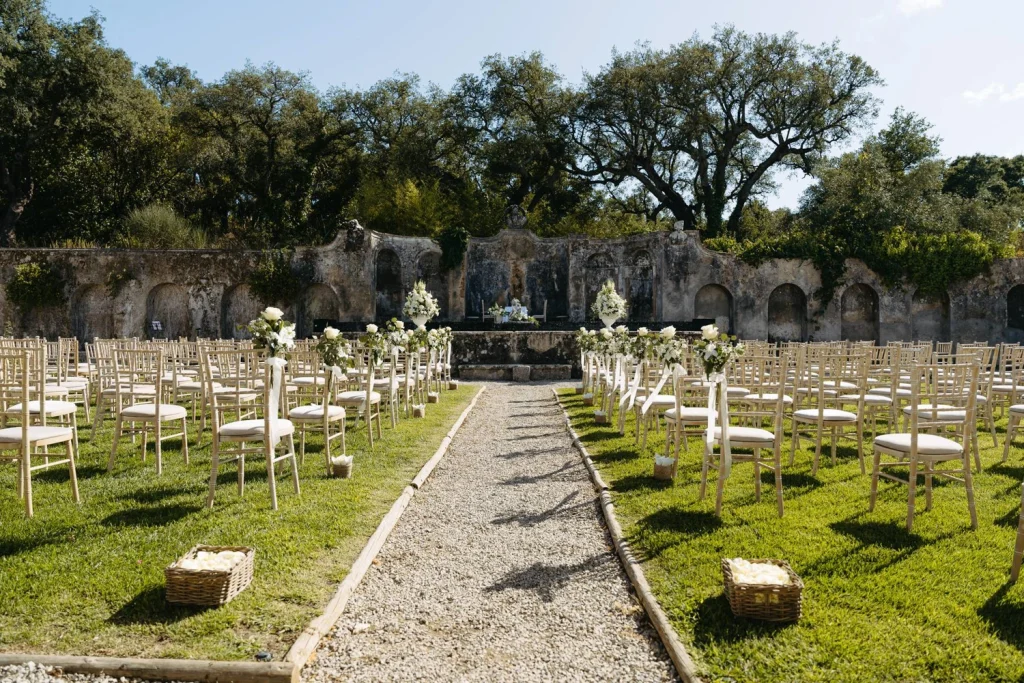 A breathtaking outdoor wedding ceremony setup with delicate white florals and elegant seating, framed by historic stone walls and lush greenery.