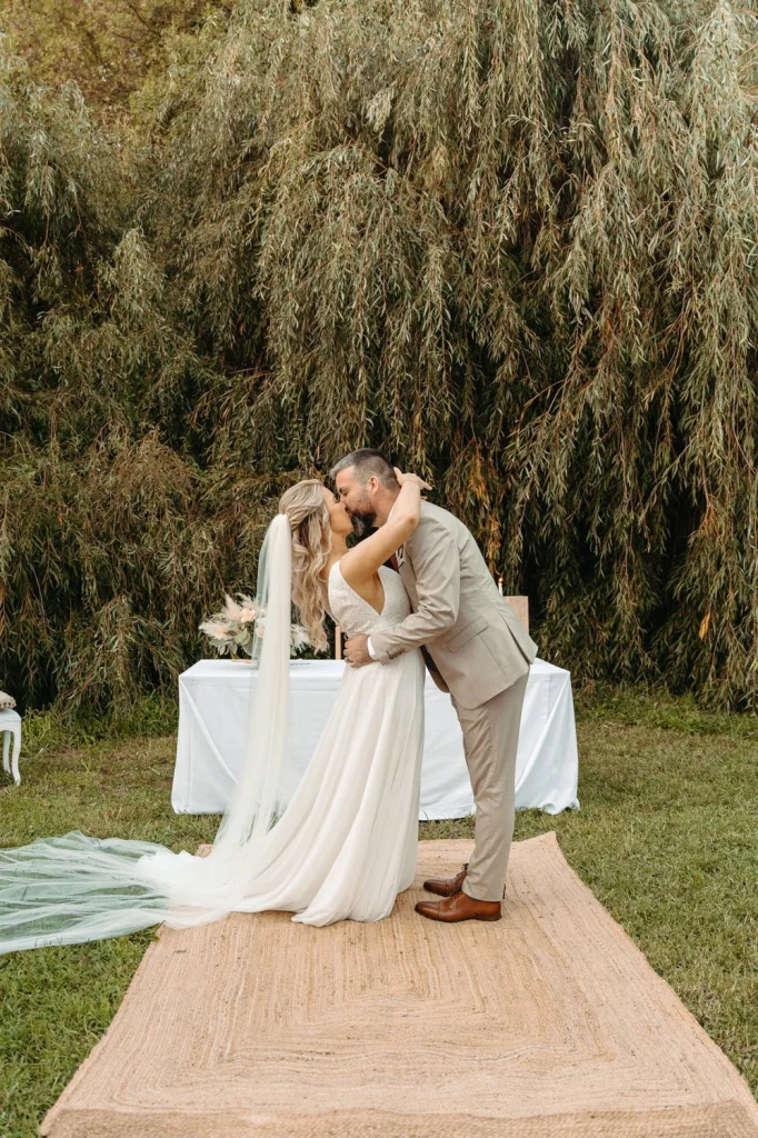 The bride and groom share their first kiss as newlyweds under a beautiful outdoor wedding setup with lush greenery in Portugal.