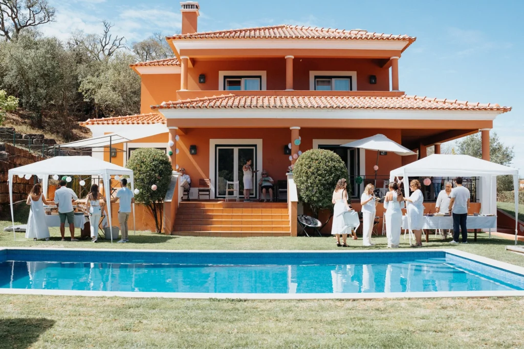 A lively outdoor celebration at a private villa in Portugal, with guests in white attire mingling by the poolside under a sunny sky.