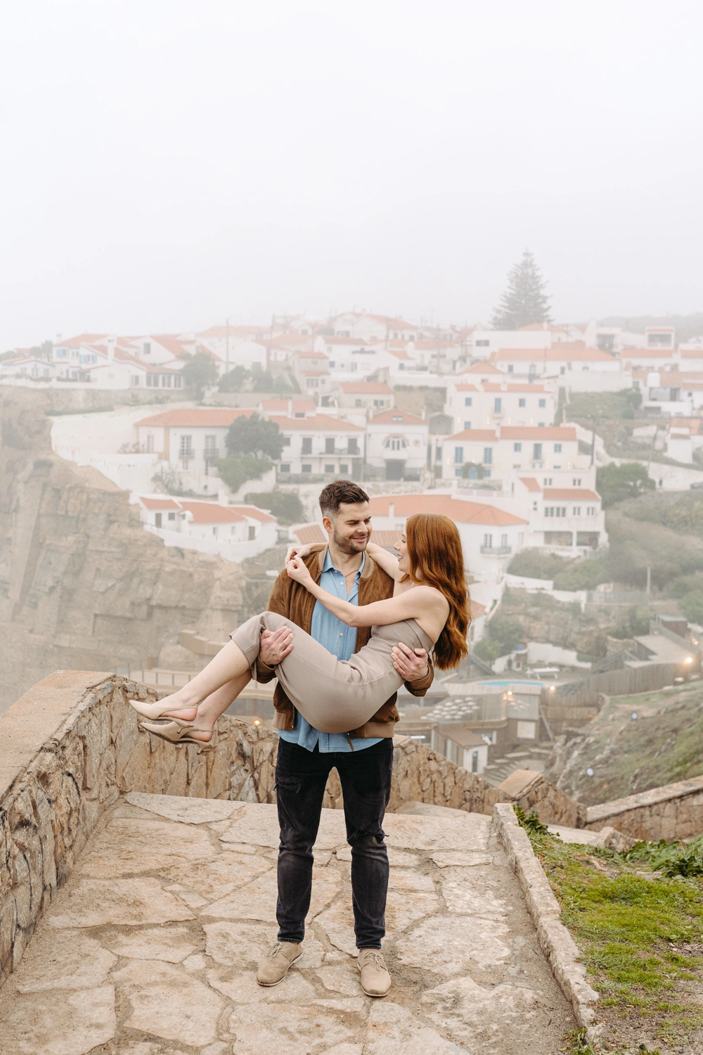 Groom-to-be playfully lifting his fiancée in his arms on a scenic cliffside overlooking a charming Portuguese village called Azenhas do Mar