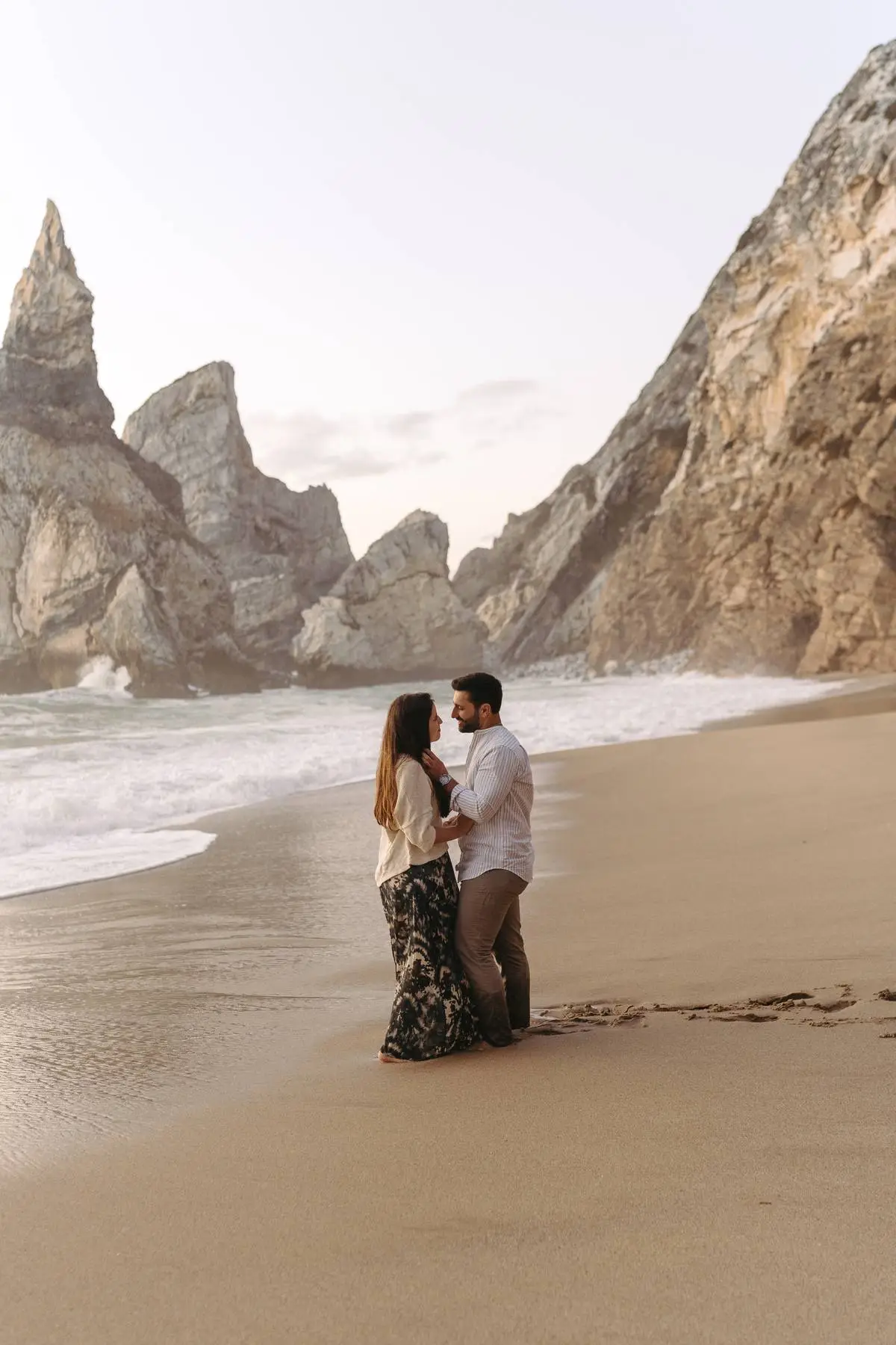 A couple shares a romantic moment on the sandy shores of Praia da Ursa, Portugal, with dramatic rock formations and ocean waves creating a breathtaking backdrop during their beach photoshoot.




