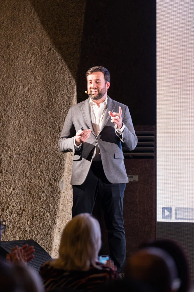 A charismatic male speaker in a gray blazer delivering a presentation at a high-end conference in Lisbon.