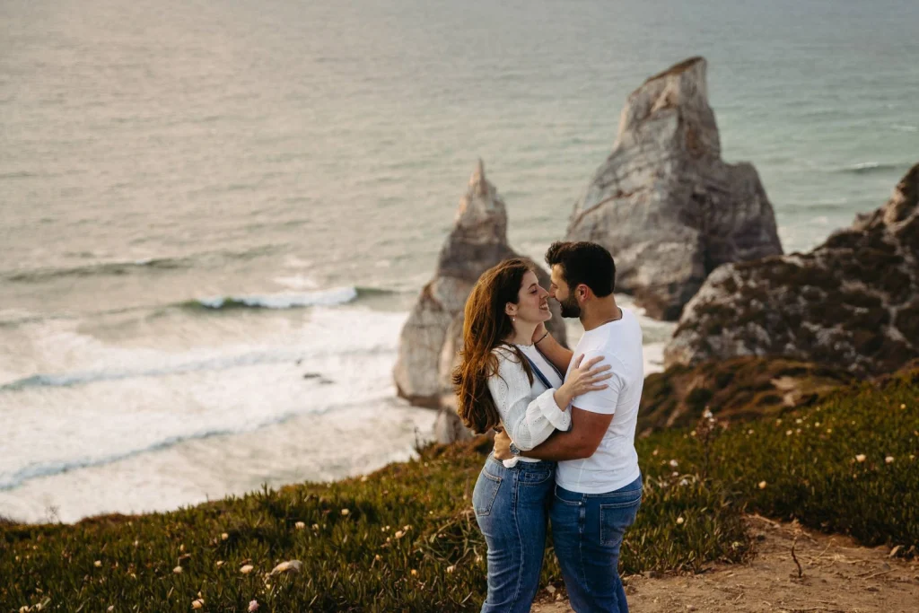 A couple sharing a romantic kiss on the beach at sunset, with ocean waves and rock formations in the background