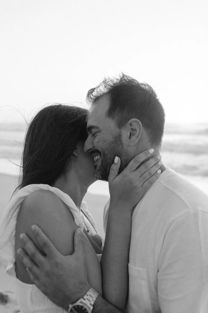 A black-and-white engagement photo capturing a couple sharing a joyful embrace on the beach.