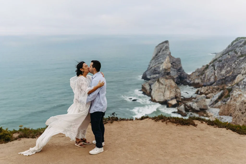 A couple kisses on a dramatic cliffside overlooking the Atlantic Ocean in Portugal, with the wind blowing around them in praia da ursa