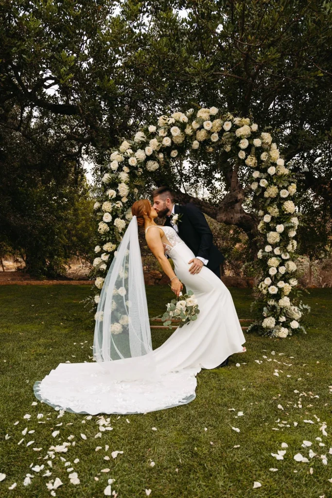 The bride and groom share a romantic kiss under a stunning floral arch, symbolizing their love and commitment.