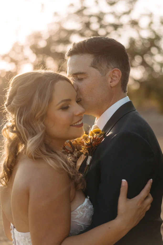 Groom kisses bride’s forehead in golden sunset light, capturing a romantic and intimate wedding moment.