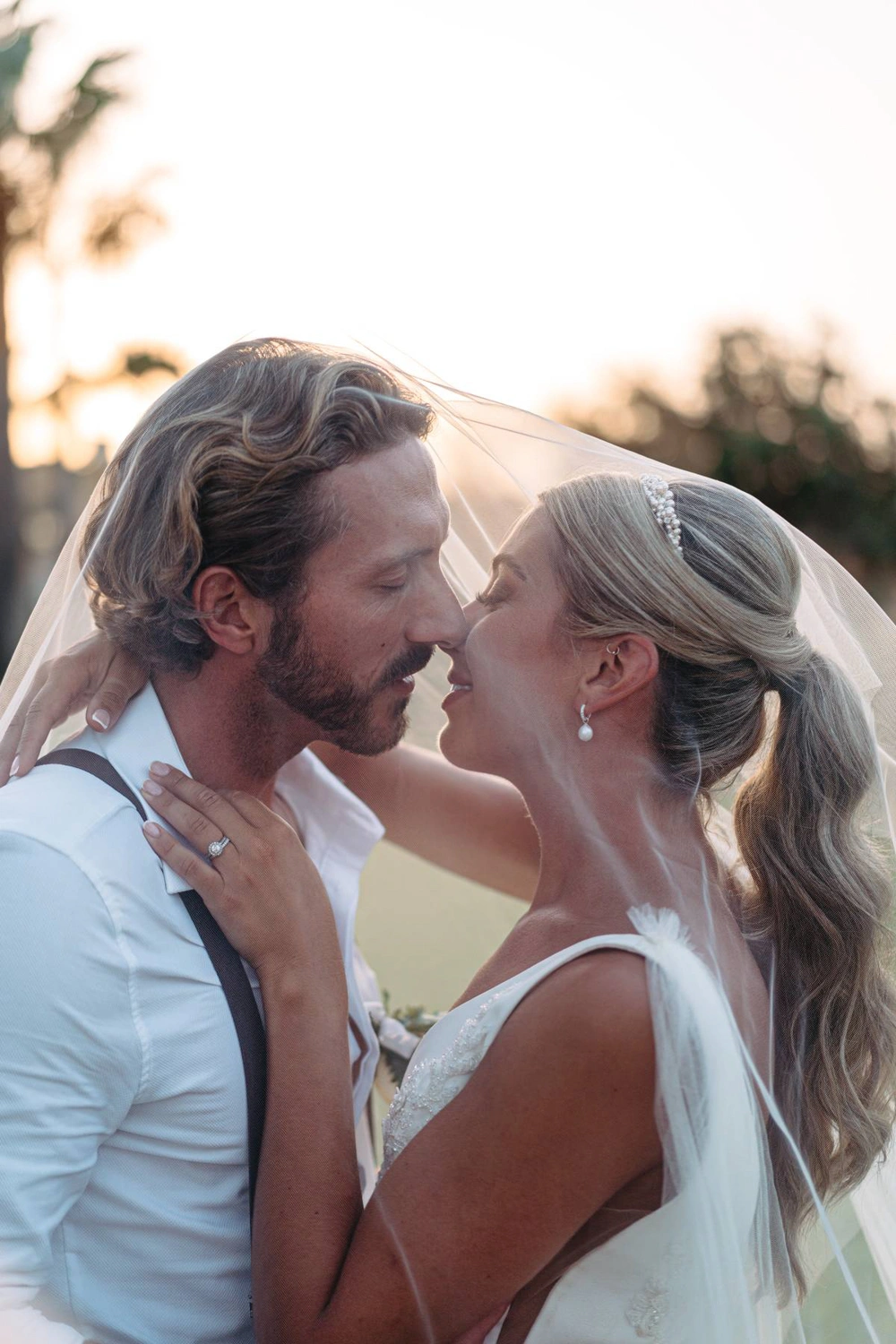 The bride and groom share a tender moment at sunset, surrounded by palm trees and glowing in the warm light of their dreamy destination wedding in Portugal.