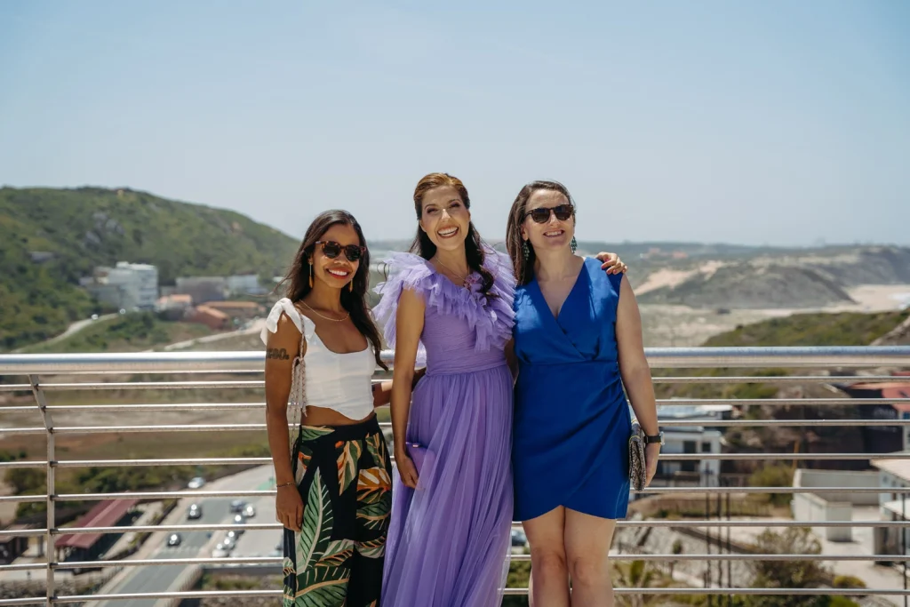 Three women pose for a joyful photo on a scenic terrace with a stunning ocean view, dressed elegantly for an event.