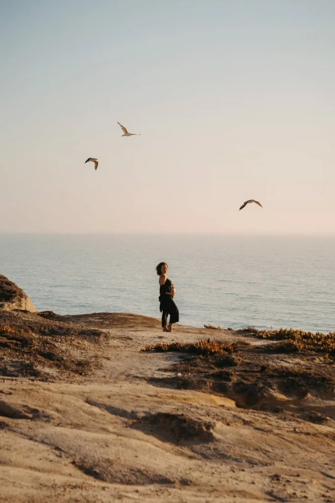 Expecting mother standing on a rocky cliffside at sunset, gazing at the ocean in an intimate maternity photoshoot
