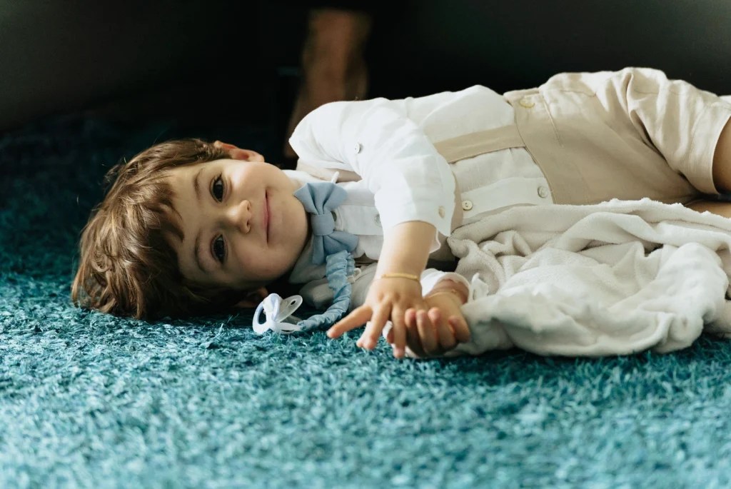 A young boy in formal attire lying on a blue carpet, smiling softly in portugal for a christening