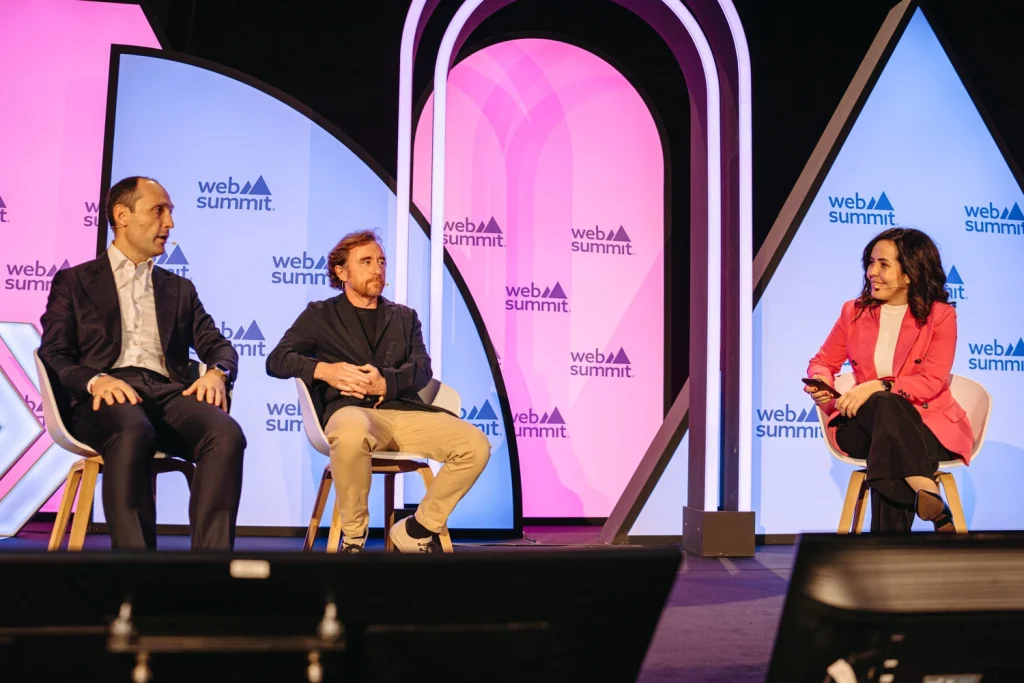 Three speakers engaged in a panel discussion on stage at Web Summit Portugal, illuminated by vibrant pink and blue lights.