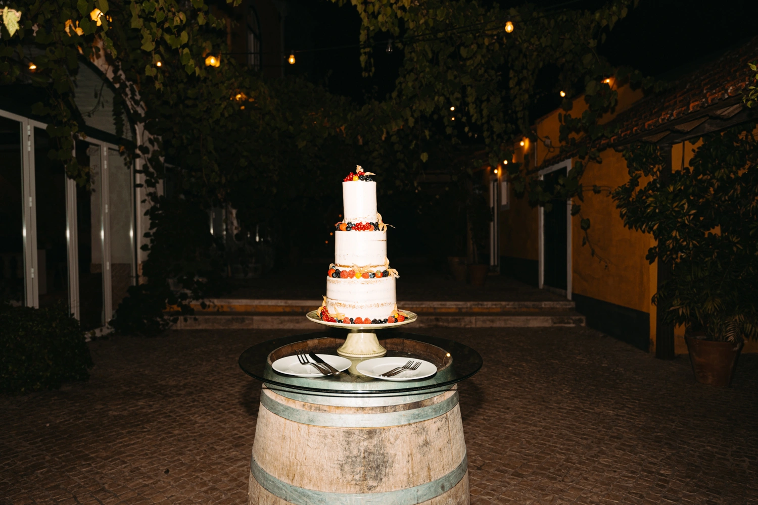 A beautiful three-tiered naked wedding cake adorned with fresh berries, set on a rustic barrel at a wedding reception.