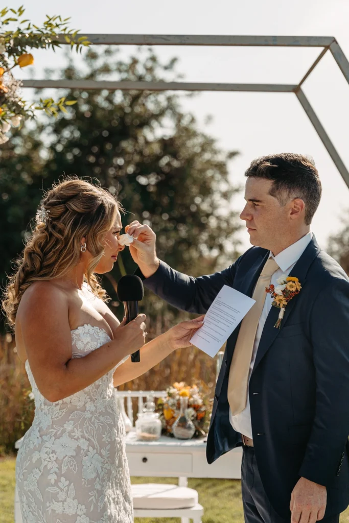 A groom gently wipes away a tear from the bride’s face as she reads her heartfelt vows during their outdoor wedding ceremony in Algarve
