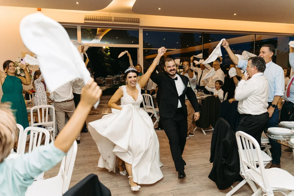 The bride and groom joyfully enter the reception, surrounded by cheering guests waving white napkins in celebration.