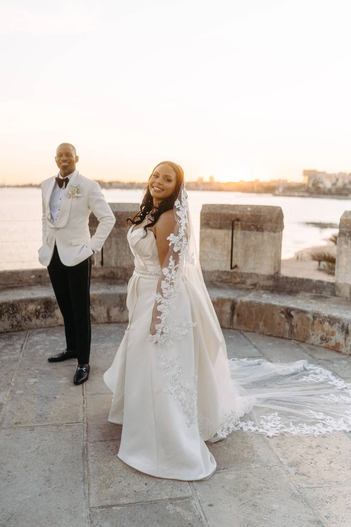 Elegant bride smiling at sunset with lace veil and long train after her Forte da Cruz wedding in Estoril Portugal.