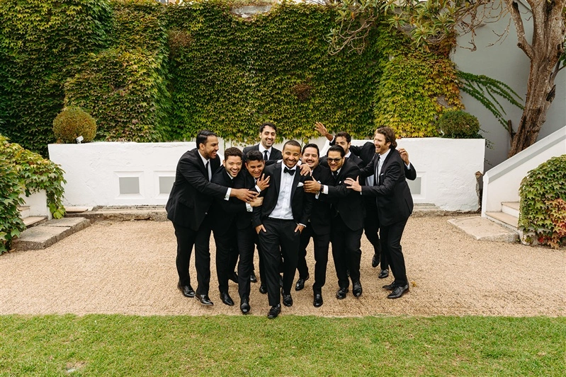 Fun and candid portrait of groom surrounded by his groomsmen in black suits at a Portugal wedding venue with ivy covered walls.