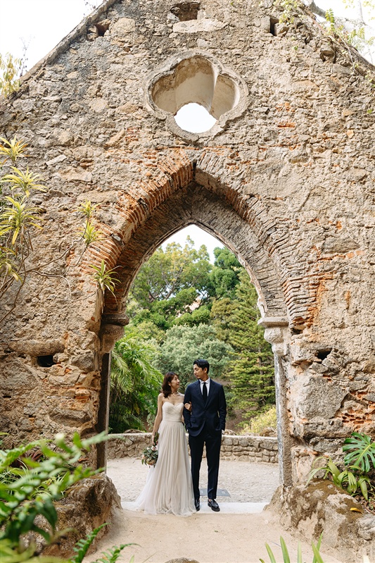 Bride and groom walking hand in hand through the arched ruins of Palacio de Monserrate in Sintra.