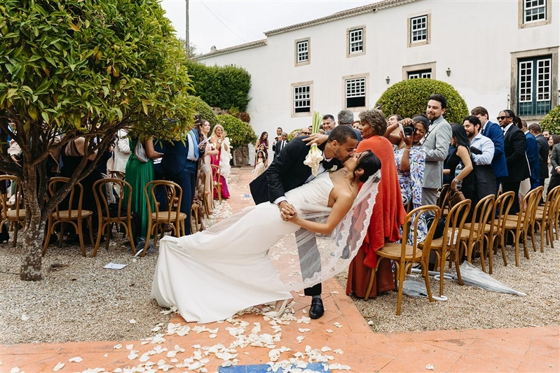 Bride and groom sharing a joyful dip kiss in the aisle after their outdoor wedding ceremony in Portugal surrounded by cheering guests.